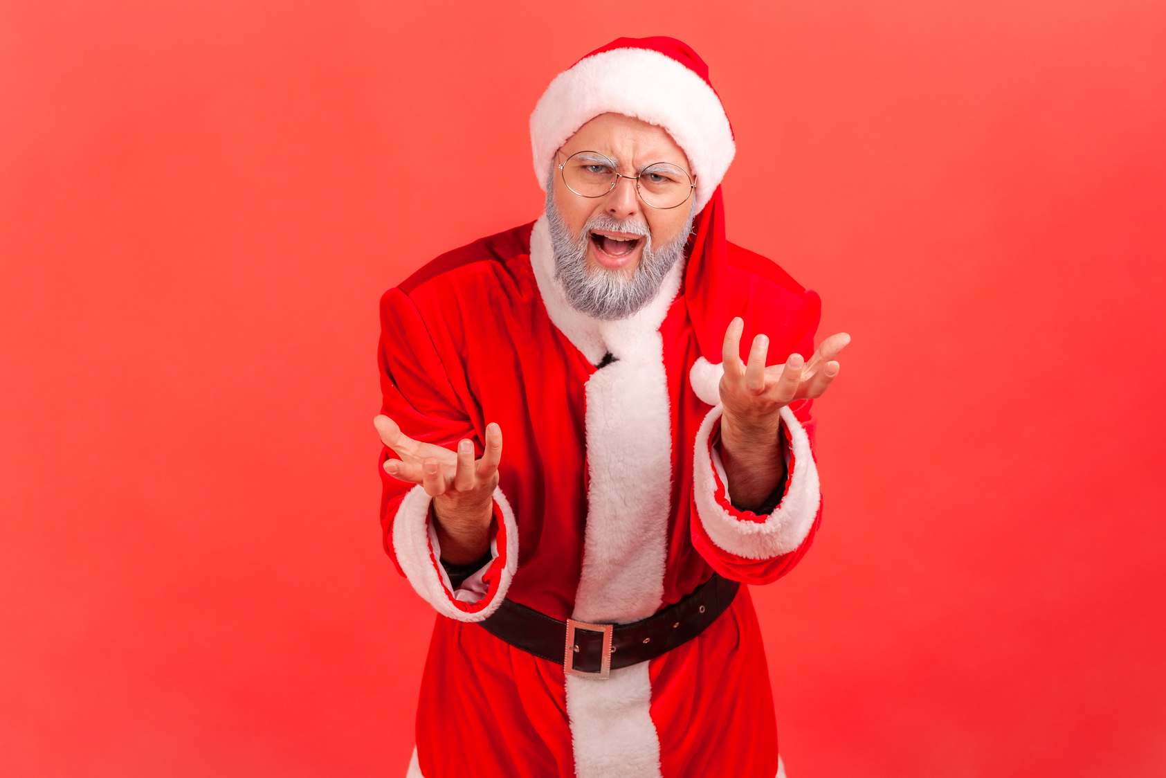 Portrait of Elderly Man with Gray Beard Wearing Santa Claus Costume Standing with Raised Hands and Indignant Aggressive Face Asking Why.
